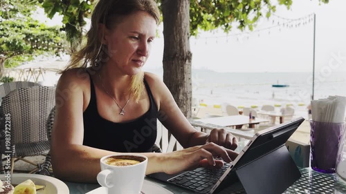 Young traveler working remotely on tablet while relaxing at coastal cafe. Digital nomad lifestyle concept combining vacation, productivity, online business and modern freelance career.