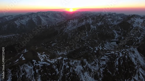 Aerial flight toward sunrise above snowy Andes Mountains with layered peaks and frozen valleys. Morning light reveals glacial lakes icy ridges vast andean wilderness beside valley.