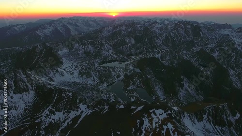 Aerial flight toward sunrise above snowy Andes Mountains with layered peaks and frozen valleys. Morning light reveals glacial lakes icy ridges vast andean wilderness beside valley.