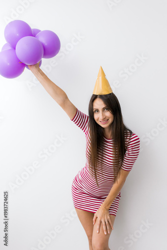 Smiling woman in a striped dress holding purple balloons while standing against a white wall. 