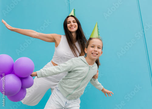 Happy mother and daughter laughing together while holding purple balloons against a bright blue background.