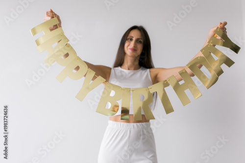 A woman dressed in white stands against a white background, holding a golden “Happy Birthday” sign. Minimalistic and elegant composition with a festive touch. 