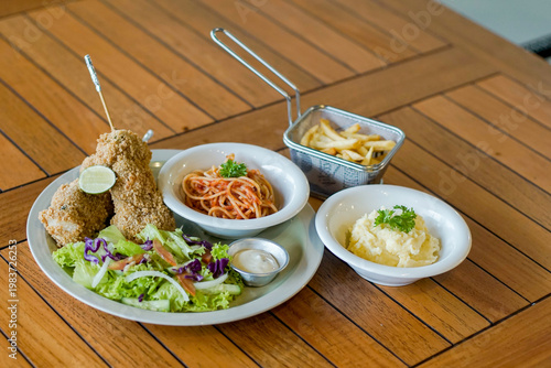 Delicious crispy fried chicken served with spaghetti, fries, mashed potatoes, and fresh salad on wooden table. Balanced comfort food meal, ideal for restaurant, menu, and food blog use.
