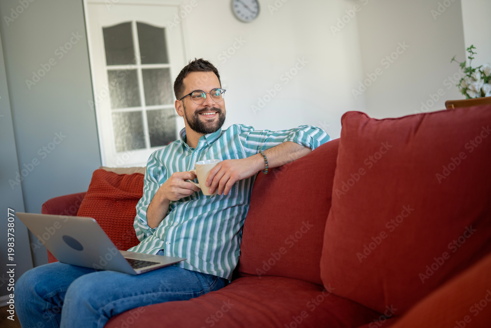 Obraz premium Smiling man relaxing on sofa, enjoying coffee and using laptop