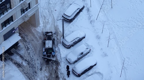 aerial top-down view of a tractor cleaning snow in a residential parking lot during winter, snow-covered ground, parked cars, winter maintenance in action