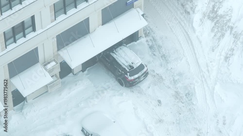 Car entering underground parking at residential building in winter, top-down aerial view with snow-covered driveway, vehicle in motion, urban winter scene