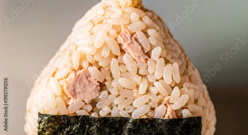 Extreme close-up of a triangular tuna mayo onigiri showing flaked tuna mixed into white rice, wrapped at the base with nori seaweed, against a soft gray background.