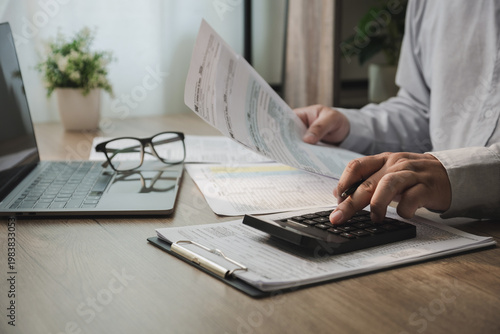 Hands writing on tax document with pen beside laptop on desk, processing financial paperwork for accounting, calculation, expense reporting, and document management in a business workflow