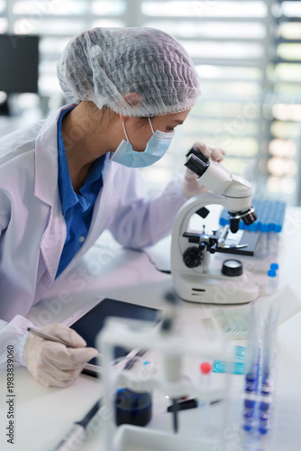Asian female scientist adult woman conducting medical research using microscope analyzing samples recording data on digital tablet in modern scientific laboratory healthcare innovation