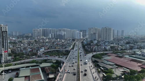 Aerial view of the roundabout leading to Vinh Tuy Bridge in Hanoi, with steady traffic and dense urban housing under approaching storm clouds.