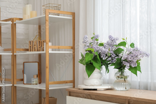 Vases with beautiful bouquets of flowers on chest of drawers and shelving unit near window