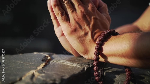 Man wearing prayer beads, hands clasped in prayer gesture on a rough stone surface, spiritual devotion, peaceful meditation, religious contemplation