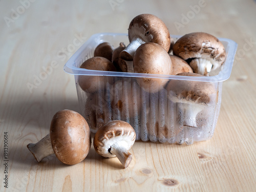 Fresh brown button mushrooms cremini mushrooms in a plastic container on a light wooden table. Natural organic food ingredient