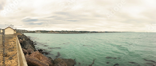 Panoramic coastal view of Friendly Bay in Oamaru with turquoise waters, rocky breakwater, and South Hill cityscape under cloudy sky - Otago, New Zealand, February 5, 2017
