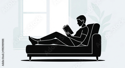 A smiling young businessman sits on a comfortable home sofa reading a book while his laptop and pc remain on the office desk in the room