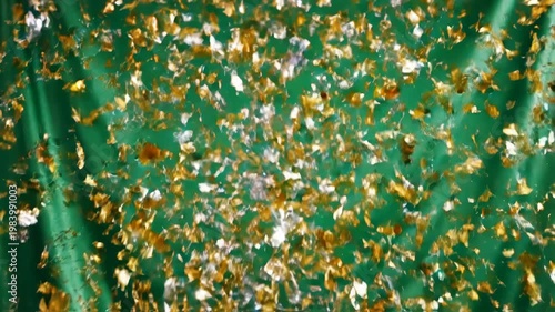 Gold and silver metallic confetti falling and scattering in front of a bright green draped fabric background with motion blur effect during a festive holiday celebration.