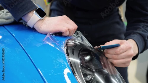 Detailer worker smoothes PPF, Paint Protection Film on blue sport car Headlight. The process of installing PPF film on the headlight of the blue car. A car wrapping specialist applies a PPF. 