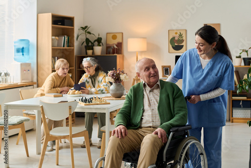 Принти на полотні Senior man with disability sitting in wheelchair while young adult caregiver assisting in nursing home