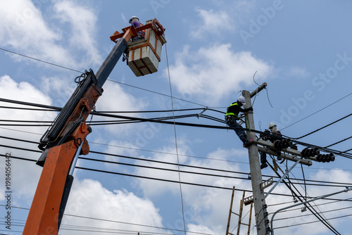 Utility workers repair and replace electrical wires on a tall pole in Thailand, using a bucket truck and safety gear to maintain a safe and operational power grid.