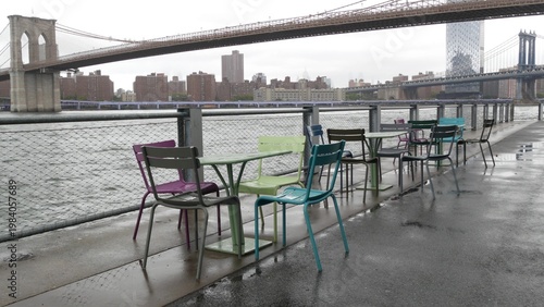 Brooklyn Bridge, Manhattan Bridge from Brooklyn Bridge park, New York City Dumbo United States. Waterfront promenade, riverside esplanade on rainy foggy day. Empty wet metallic chairs, tables by water