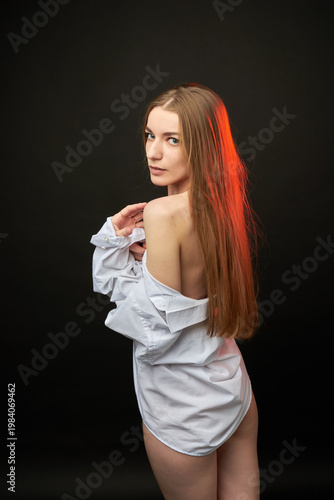 A young woman poses in a white off-the-shoulder shirt on a black background. The soft lighting highlights her relaxed and confident expression. She is wearing minimal underwear, which adds to her sens