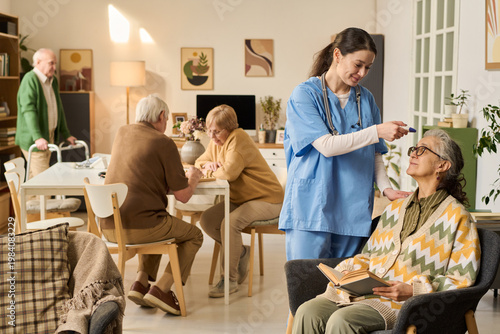 Fotografija Senior woman receiving temperature check from female nurse while reading in care home, with elderly residents socializing behind