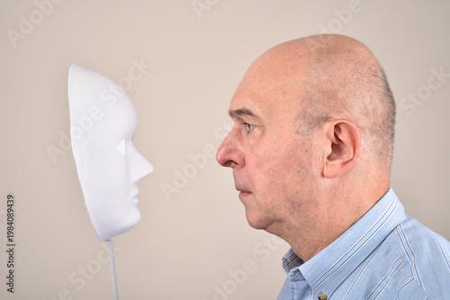 Frowning middle-aged man looking at the white mask and trying to understand dissociative identity disorder. 