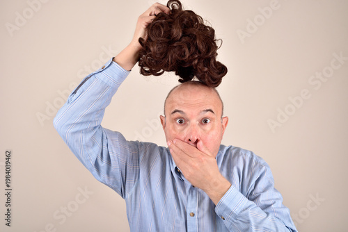 Wide-eyed surprised and astonished middle-aged bald man with a hand over his mouth taking off his wig. Health or aging problem concept.
