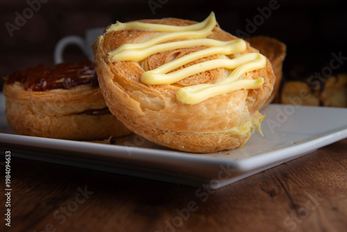 Reposteria casera, pan de hojaldre sobre un plato blanco para disfrutar en familia con cafe o leche, cubierta de crema pastelera de vainilla, vista lateral macro.