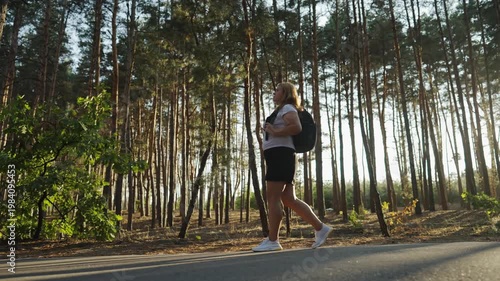 Woman with backpack walking on paved road through pine forest, sunlight through trees, active outdoor recreation in nature.