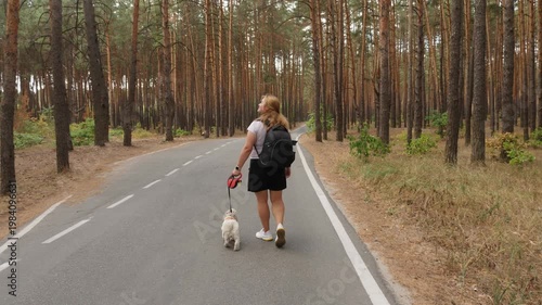 Young woman with black backpack and small white dog on leash walking along asphalt road through pine forest. Back view, active outdoor recreation, traveling with pet in woodland.