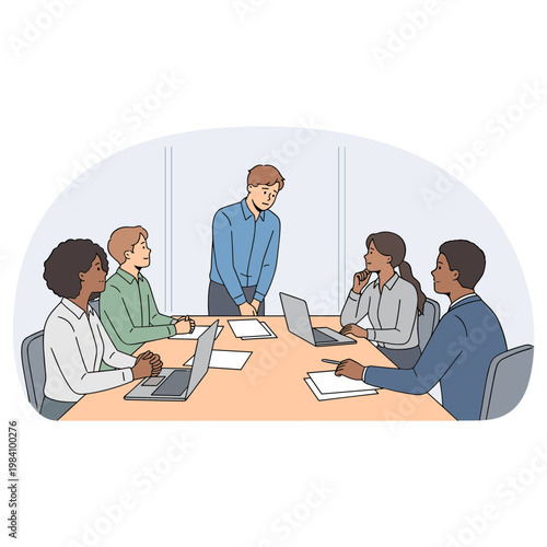 A diverse group of people are sitting around a conference table with laptops while one man stands presenting.
