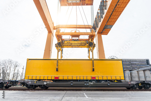 Gantry crane lowering a yellow semi-trailer onto a flatbed freight wagon at an intermodal rail terminal, professional cargo loading for efficient rail and road transport logistics