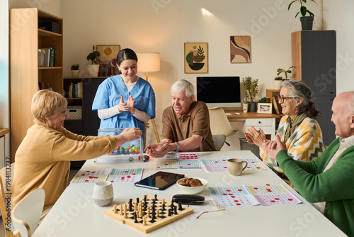 Slika na platnu Senior group playing bingo with female caregiver, interacting around activity table in care facility