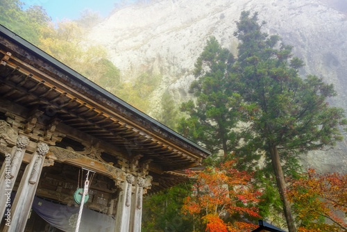 Ornate temple hall on Shikoku Henro beneath misty cliff with autumn foliage in Japan