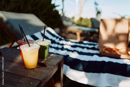 Konstfotografi Outdoors picnic scene with a wooden table, colorful drinks, a striped blanket, and warm sun, capturing casual relaxation and a bright summer lifestyle in a simple, candid moment