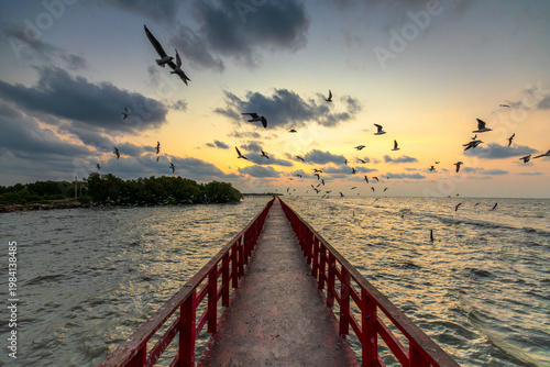 Fotografia Red wooden bridge over the sea at sunset with seagulls flying,A perspective view of a long red wooden bridge stretching into the sea during a dramatic golden sunset
