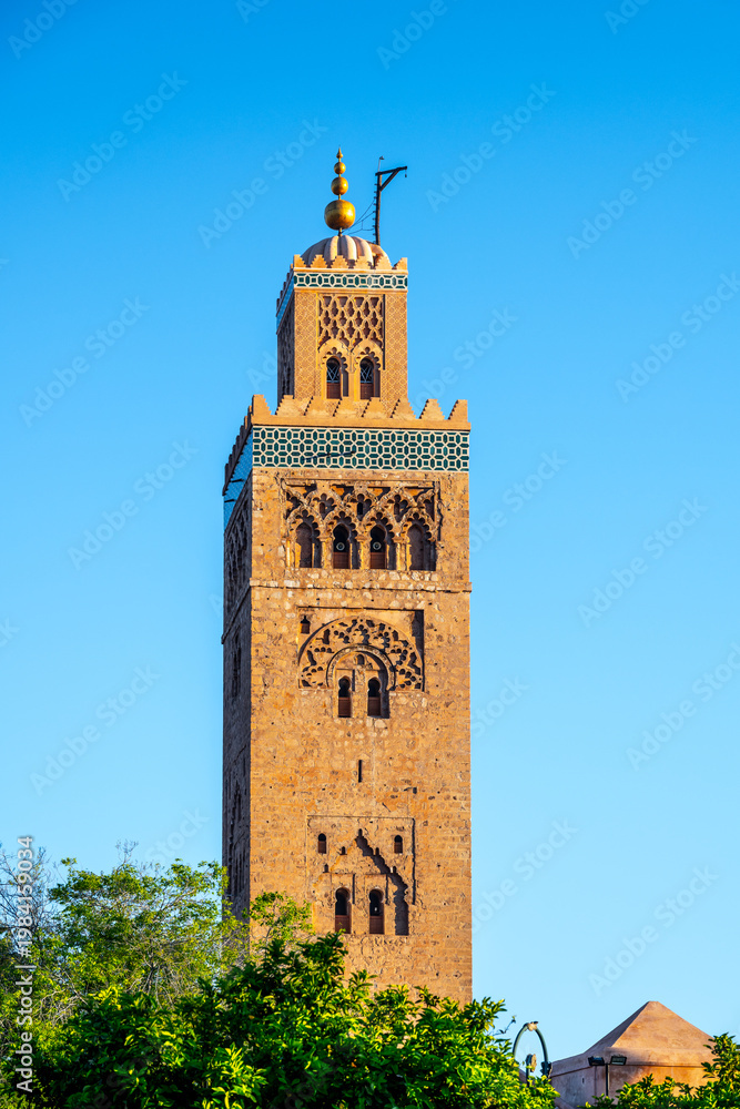 Naklejka premium The tower of Kutubiyya Mosque stands tall in Marrakesh, Morocco. The clear blue sky forms a backdrop as the sun shines on this historical site. Visitors may be nearby, enjoying the scene.
