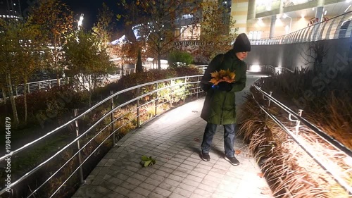 Young woman walking in illuminated autumn city park at night with bouquet of fallen leaves