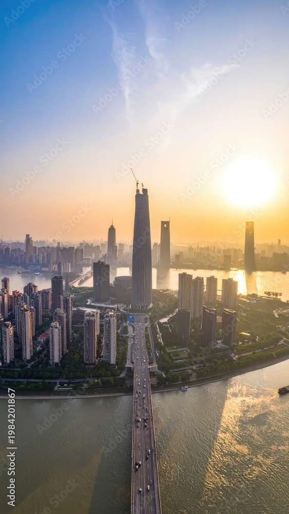 Fototapeta premium An aerial view captures a sprawling cityscape bathed in the warm glow of a setting sun, showcasing modern architecture and a prominent bridge connecting the urban landscape.