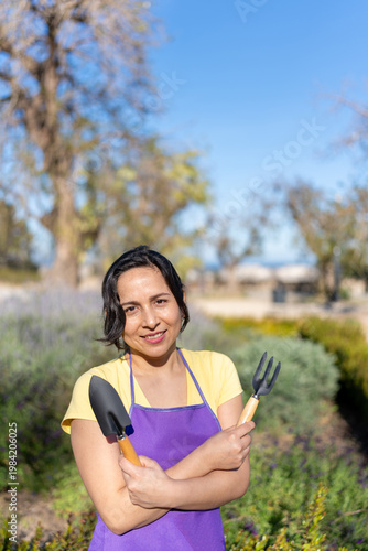Latin woman wearing an apron, smiling while holding gardening tools outdoors