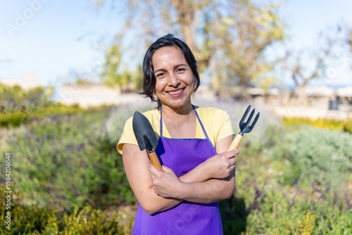 Happy Latin woman gardener smiling, holding gardening tools in an outdoor field