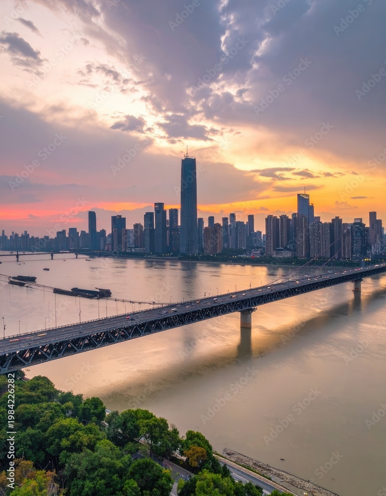 Obraz premium A stunning aerial view captures a modern cityscape illuminated by a vibrant sunset, with a bridge gracefully spanning a wide river.