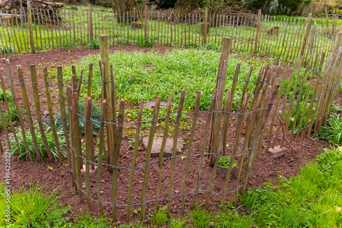 Rustic garden enclosure crafted from wooden stakes and wire, surrounded by lush green grass and vibrant plant life