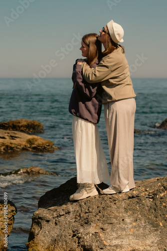 Mother tenderly hugs teenage daughter standing on large rock on seashore in sunny day