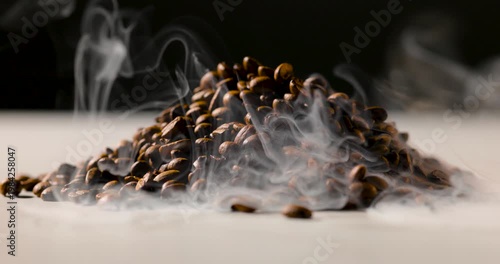 A pile of roasted coffee beans emits gentle steam, creating a rich, aromatic atmosphere against a dark background. Shallow depth of field highlights texture and warmth
