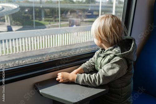 Wallpaper Mural Blond White Child Watching Passing Highway, Hands On Tray Table, Blue Seat, Urban Overpass And Parked Cars Outside Window, Daytime Light, Relaxed Travel Vibe, Anticipation For Family Destination, Torontodigital.ca