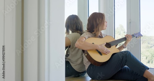 African American woman sitting on hardwood floor, with guitar, strumming beside windows, singing