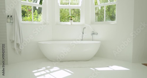 Freestanding oval bathtub sitting centered under three-pane windows catching sunlit floor glow