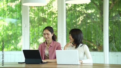 Two young women smiling and collaborating on a laptop in a bright modern workspace, representing teamwork, productivity, and a positive working atmosphere.